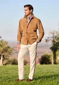 Veste en daim beige avec un col montant, fermeture à boutons et deux poches avant. Associée à une chemise à rayures de couleur claire et un pantalon crème.