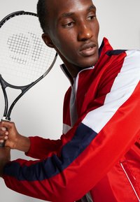 Chaqueta deportiva roja y azul marino con tejido texturizado, con mangas en bloque de color. Sostiene una raqueta de tenis negra con una malla.