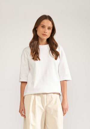 Pitkähiuksinen ruskeatukkainen nainen, jolla on valkoinen ylisuurikokoinen lyhythihainen paita ja beiget leveälahkeiset housut, seisoo tasaisen taustan edessä.