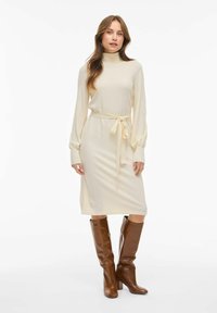 Robe crème à manches longues avec col montant, taille ceinturée et texture douce. Associée à des bottes brunes à talons montantes jusqu'aux genoux.