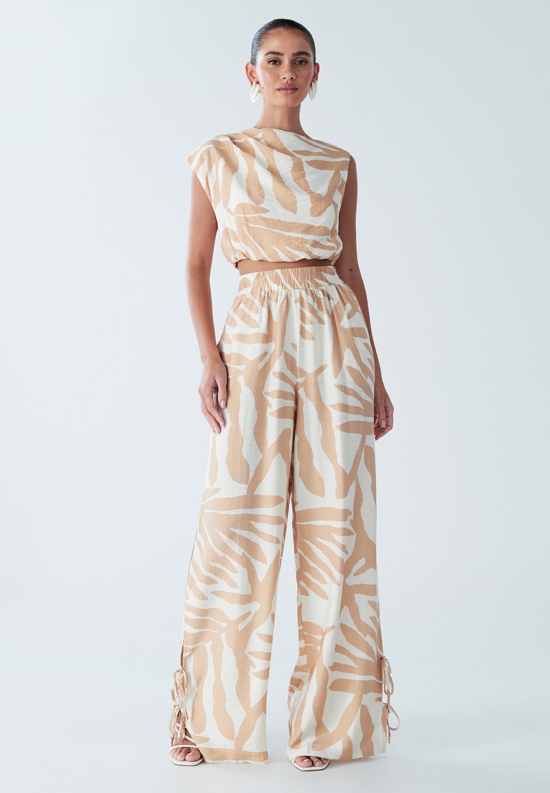 Beige dvoudílný outfit sestávající z kráceného topu s krátkými rukávy a kalhot širokého střihu, zdobený bílým abstraktním vzorem a postranními tkaničkami.
