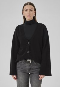 Cardigan negru dintr-un material moale, cu un decolteu adânc în V, două nasturi și mâneci largi. Purtați peste un pulover negru cu gât înalt și blugi gri.
