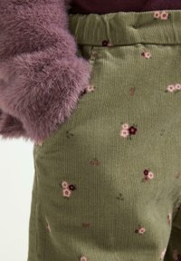 Pantaloni in corduroy verde con un motivo floreale in rosa e bordeaux, con una morbida manica pelosa viola che si sovrappone alla cintura.