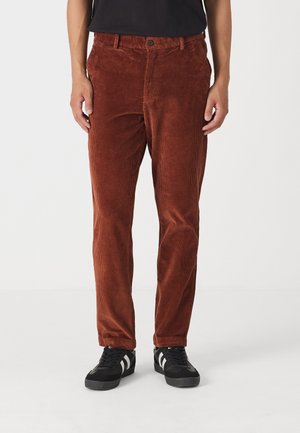 Rust farvede kordfløjsbukser med lodrette ribber, der har en knaplukning, bæltestropper og forlommer. Båret sammen med sorte sneakers.