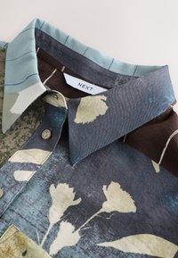 Chemise à manches courtes présentant un motif floral aux teintes vertes, bleues et marron, un col à boutons, et une texture de tissu lisse.