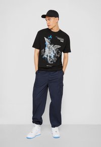Schwarzes Grafik-T-Shirt mit Engel-Design in Blau und Weiß, kombiniert mit dunkelblauen Cargo-Hosen und weißen Sneakern. Modell trägt eine schwarze Mütze.