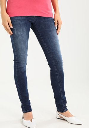 Osoba ubrana w ciemnoniebieskie obcisłe dżinsy, różowy top i białe baleriny na spiczastym nosku, stojąca ze swobodnie opuszczonymi rękami.