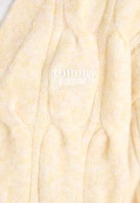 Hele kollase lõngaga kootud sukkel, millel on kootud muster ja pehme tekstuur. "Tommy Jeans" logo on riidele valgete tähtedega tikitud.