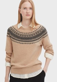 Beige Strickpullover mit einem schwarzen gemusterten Einsatz, rundem Halsausschnitt und geripptem Saum. Getragen über einem weißen Hemd mit Kragen. Weiche Textur.