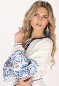 Witte blouse met blauwe geborduurde patronen, ingeknoopte taille; heeft lange mouwen met gerimpelde manchetten, aangevuld met blauwe druppel oorbellen.