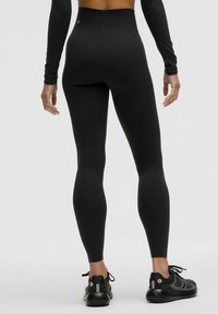 Sorte højtaljede leggings lavet af strækbart materiale, tætsiddende design der viser en glat tekstur. Bæres med sorte træningssko.