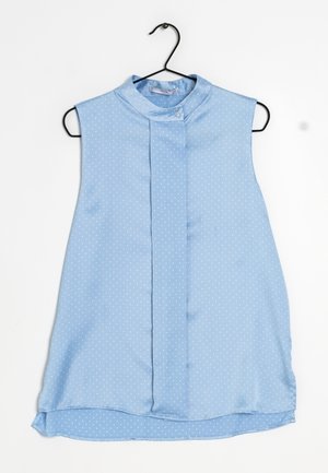 Blouse sans manches bleu clair à petits pois blancs, col montant avec bouton, suspendue sur un cintre noir.