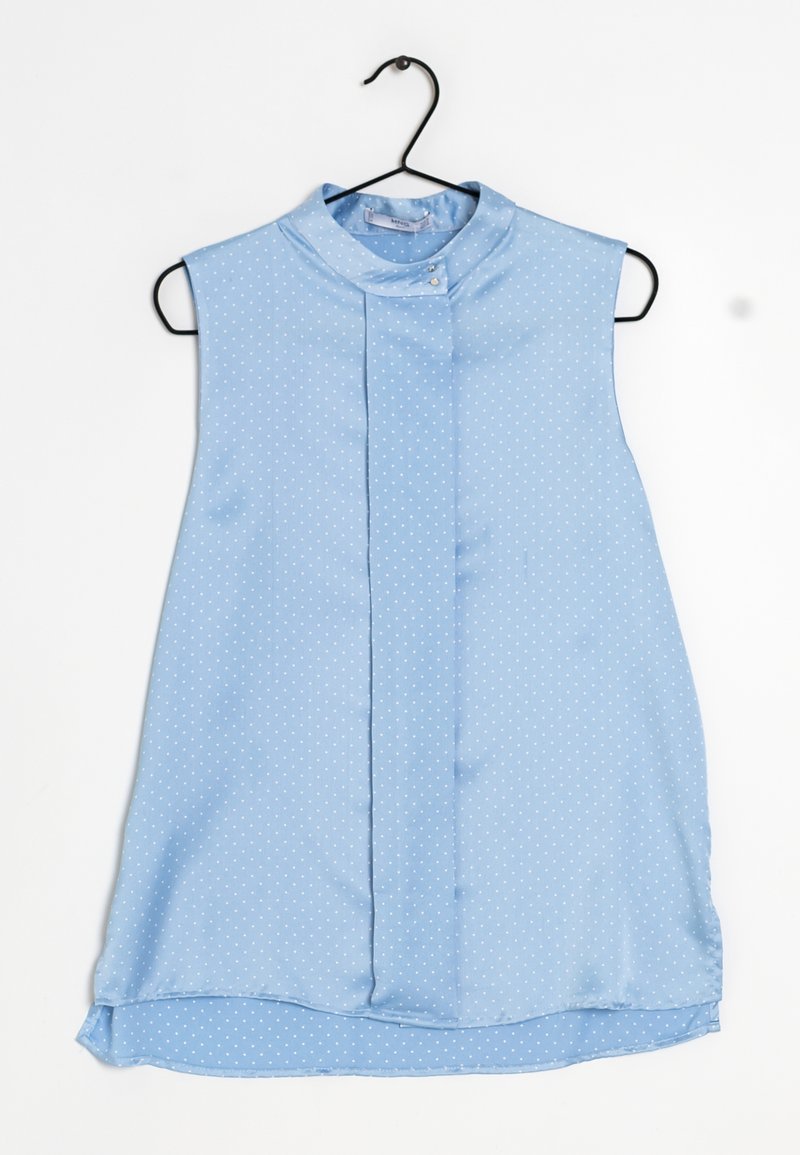Blouse sans manches bleu clair à petits pois blancs, col montant avec bouton, suspendue sur un cintre noir.