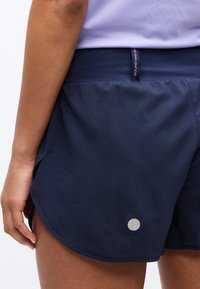 Κοντινό πλάνο ενός ατόμου που φοράει σκούρο μπλε αθλητικό σορτς με μια μικρή στρογγυλή ανακλαστική λεπτομέρεια κοντά στο στρίφωμα και μια μαύρη ετικέτα μέσης.