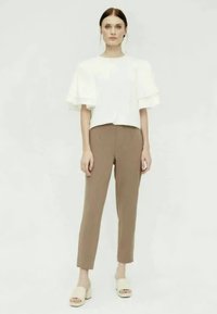 Witte korte mouwen top met rimpeldetails en bruine op maat gemaakte broeken. Het model draagt beige instapsandalen, geplaatst tegen een neutrale achtergrond.