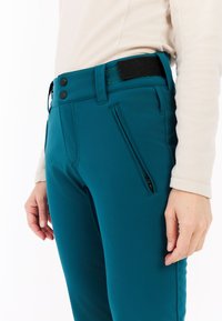 Mėlynos elastinės kelnės pasižymi siauru siluetu, užtrauktuku užsegamu kišeniu ir juodu knopku. Juosmens srityje yra Velcro tvirtinimo juosta.