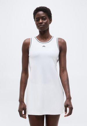 Mujer vistiendo un vestido deportivo blanco sin mangas con ribetes negros y un pequeño logo negro en el pecho, de pie contra un fondo liso.