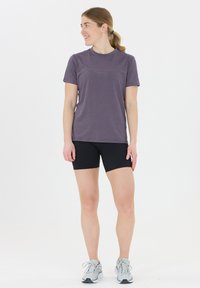 Lila kortärmad träningströja med rund halsringning, tillsammans med svarta shorts. Modellen bär grå träningsskor. Enkla, funktionella design.