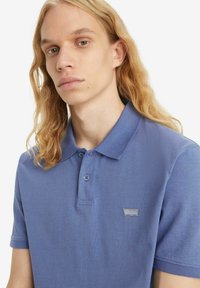 Polo shirt blu in cotone, con colletto, due bottoni e una piccola patch con logo sul lato sinistro. Maniche corte con una texture liscia.