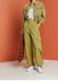 Pantalon cargo vert olive à jambes larges avec de grandes poches latérales, associé à une chemise ample de couleur claire et des baskets blanches. Sac tressé marron.