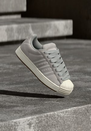 Sivá kožená teniska s plochými šnúrkami, gumovou špičkou a jemným dizajnom s tromi pásmi. Má svetlosivú podšívku a gumovú podrážku.