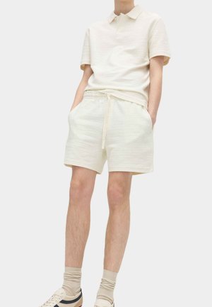 Persona vistiendo una camisa polo de manga corta en crema con textura y pantalones cortos a juego con cordón, calcetas beige y zapatillas blancas.