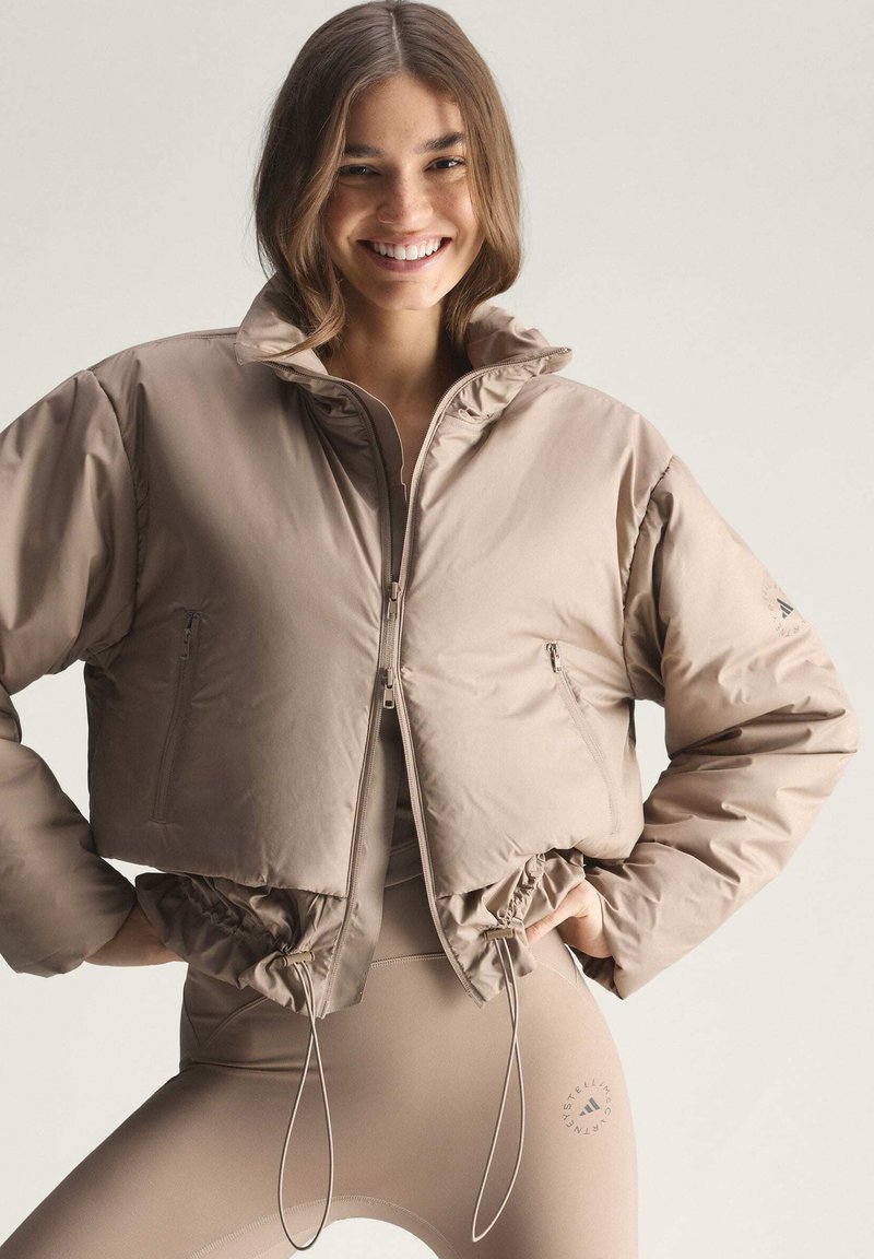 Smilende kvinde iført beige cropped dunjakke og matchende leggings med logo, stående med hænderne på hoften mod en lys baggrund.