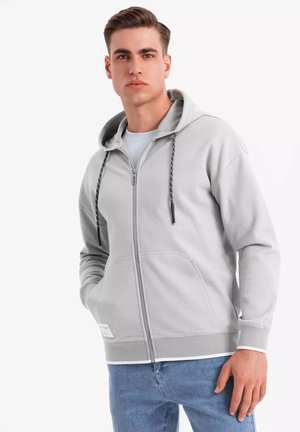 Sweat à zip gris clair avec une finition texturée, capuche à cordon de serrage, poches latérales et poignets côtelés ; porté sur une chemise bleu clair.