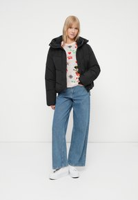 Sort pufferjakke med en cropped design og hætte, båret over en mønstret cremefarvet sweater. Sammen med vide blå jeans og hvide sneakers.