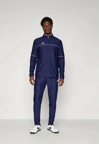 Vīrietis stāv armijas zila krāsā pilna garuma sporta uzvalkā ar baltām līnijām un baltām kedām uz vienkārša gaiša fona.