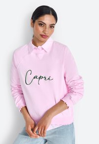 Felpa rosa con testo ricamato "Capri" verde, colletto a coste e polsini ondulati. Tessuto liscio con una vestibilità comoda, indossata sopra jeans chiari.