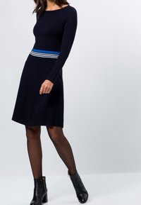 Marinblå stickad klänning med långärmar och en åtsittande överdel, med ett horisontellt flerfärgat randigt midjeband och en utställd kjol.