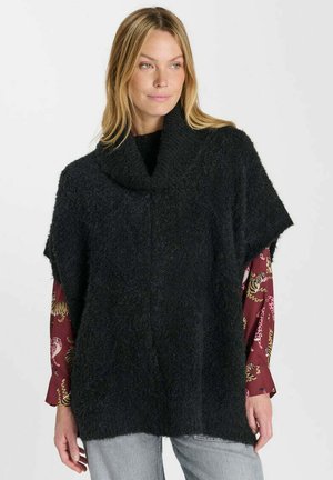 Donna che indossa un poncho di lana nero largo sopra una camicia rossa a maniche lunghe con stampe animali e pantaloni grigio chiaro su uno sfondo uniforme.