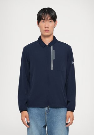 Marineblå zip-jakke med krave, der har en reflekterende lynlås og lomme-detaljer, lavet af glat stof med lange ærmer.