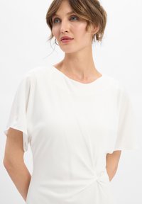 Femme aux cheveux châtains clairs courts portant un haut blanc à manches courtes avec un petit détail noué à la taille.