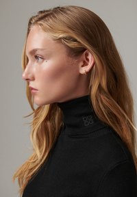 Jeune femme aux longs cheveux blonds portant un pull à col roulé côtelé noir, vue de profil sur un fond uni.