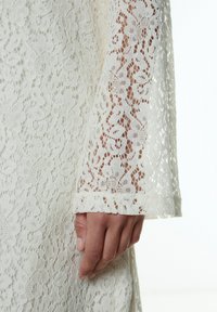 Weiße Spitzendressärmel mit floralen Mustern, transparenter Textur und gewelltem Saum. Der glatte Stoff fällt sanft über die Hand.