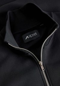 Chaqueta negra con cremallera y cuello alto, hecha de tela lisa. Cuenta con una cremallera plateada y una etiqueta que dice "aprel" dentro del cuello.