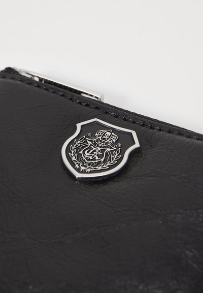 Svart läderplånbok med en silverfärgad sköld-emblem med detaljerad gravyr och en slät, strukturerad yta. Dragkedjestängning synlig.