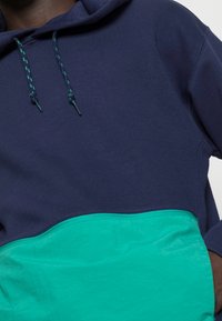 Man i en marinblå och teal färgblock hoodie med snoddar, hand i fickan, mot en neutral ljus bakgrund.