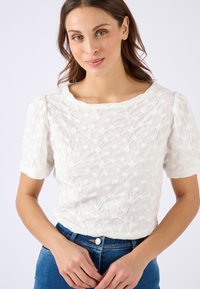 Witte korte mouwen top met een textuurpatroon van bloemen, ronde halslijn en puff mouwen, gedragen met blauwe jeans. Focus op materiaaleigenschappen en ontwerpelementen.