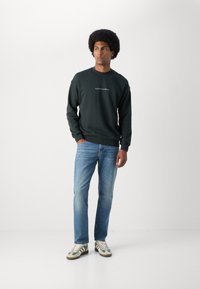 Sweatshirt vert foncé avec un logo blanc sur la poitrine, associé à un jean bleu clair et des baskets rayées vertes. Texture lisse, design décontracté.