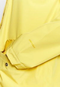 Manche de veste jaune imperméable avec le texte "DRYVENT" visible près du poignet et un bouton-pression métallique sur le corps de la veste.