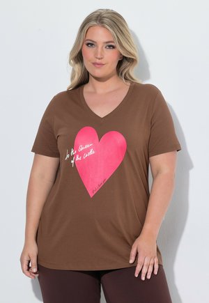Blonde Frau trägt ein braunes V-Ausschnitt-Shirt mit einem großen pinken Herz und weißem kursivem Text auf der Vorderseite, steht vor einem hellen Hintergrund.