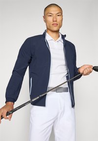 Navy zip-up jacket with textured fabric, white polo shirt, white pants, and a gray belt. Holding a golf club, featuring a sleek design.