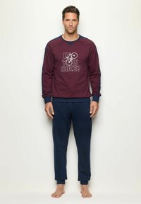 Sudadera color marrón con mangas azules que presenta el texto y gráfico "¿Andas en bici?", combinada con pantalones de chándal navy. El tejido parece suave y texturizado.