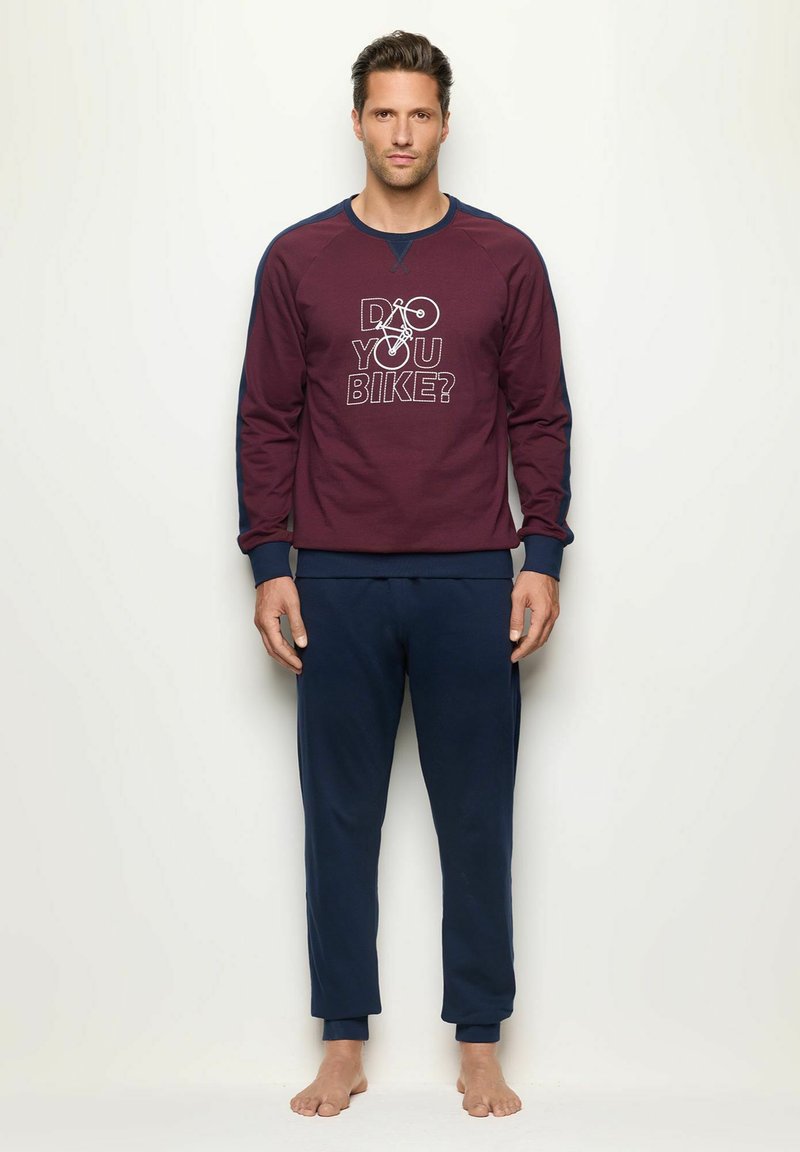 Sudadera color marrón con mangas azules que presenta el texto y gráfico "¿Andas en bici?", combinada con pantalones de chándal navy. El tejido parece suave y texturizado.
