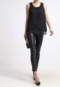 Top nero senza maniche con design drappeggiato, abbinato a pantaloni in pelle nera attillati e tacchi a punta neri, mentre tiene una piccola pochette nera.