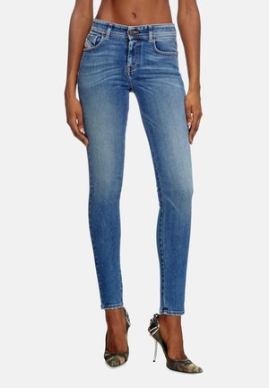 Slim-fit blå jeans båret af en stående person parret med høje hæle med camouflage-mønster mod en ensfarvet baggrund.