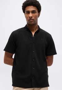 Homme aux cheveux courts et bouclés portant une chemise noire à manches courtes boutonnée, debout devant un fond clair uni.